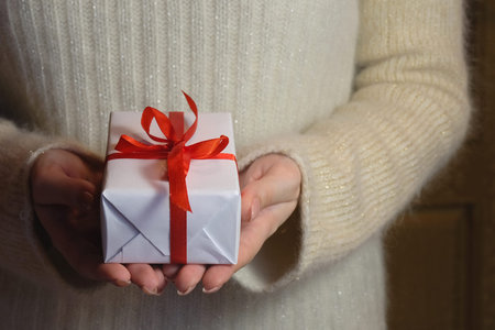female hand holding a gift box with white and red ribbon for Christmas and New Year or greeting...