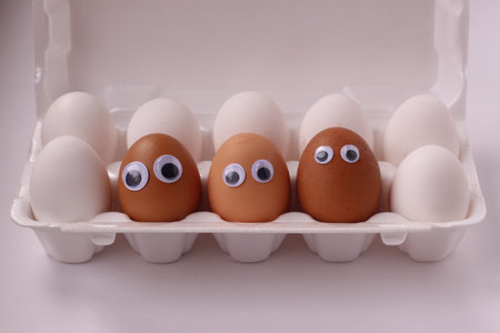 three brown eggs with eyes in a box of white eggs.