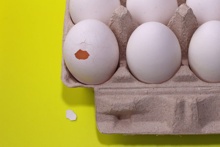 Broken egg in box with white raw eggs on yellow background.