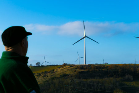 A man in a cap looks at the spinning wind turbines against the blue sky. The concept of an...