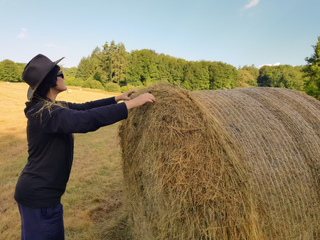 A farmer woman in a hat stands near a stack of fresh hay after harvesting wheat.の写真素材