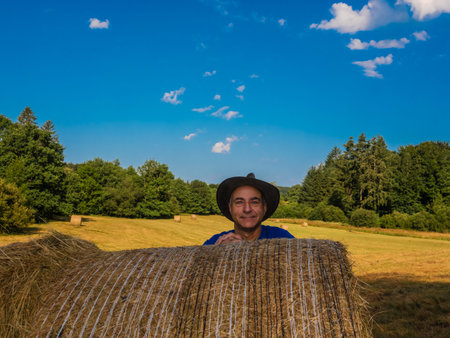 Man of a man in a hat behind a haystack in the field.