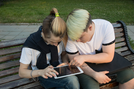 Two  friends sitting on bench  in park  and watching tablet. Have fun on line playing gameの写真素材