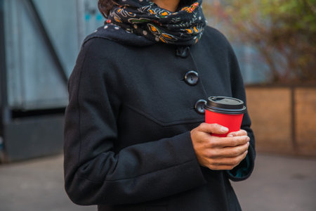 freezing young lady in stylish black coat with black scarf holding a cup of coffee