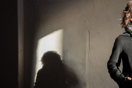 The black shadow on the white wall of young lady in black stylish coat with black scarf enjoying the sun rays at sunset in the white tunnel in the cold autumn dayの写真素材