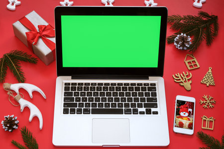 Laptop with green screen, mobile phone and Christmas decorations on red background.Concept: selection gifts, online shopping, Christmas sales, seasonal advertising.Mockup postcard, holiday invitation.の写真素材