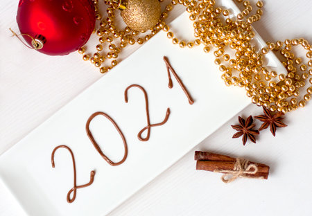 Christmas card 2021 made of chocolate on a white square plate. Christmas toys and Christmas beads.