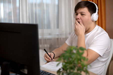 A young man in a white t-shirt and headphones is sitting at a table, listening to an online lesson and yawning. The idea of remote education education during the coronavirus pandemicの写真素材