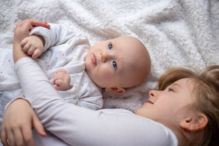 Happy children, toddler and older sister, hugging at home on a white blanket, smiling, shot from...