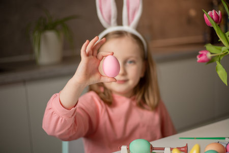 A cute little girl in rabbit ears is preparing painted Easter eggs for Easter sitting at the kitchen...
