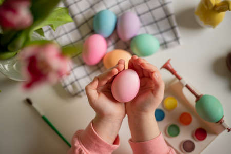 easter egg in the hands of a little girl over a table with painted eggs, watercolors and tulips.の写真素材