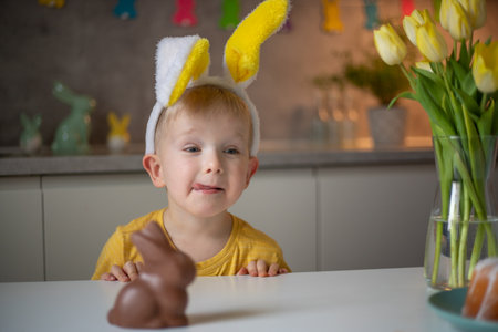 A cute little boy wearing bunny ears on Easter day wants to eat a chocolate easter bunny. A child plays egg hunt for Easter. Charming child celebrates Easter at home.の写真素材