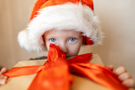 Young child in a Santa hat excitedly peeking at a beautifully wrapped gift during the festive Christmas seasonの写真素材