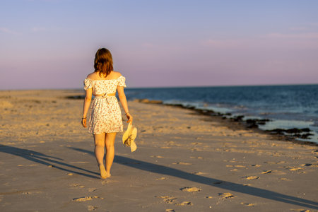 Young woman walks barefoot on the beach during sunset, holding a light sweater while exploring the shorelineの写真素材