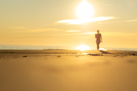 Person walks along the beach at sunset with sun reflecting on the waterの写真素材