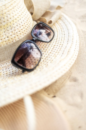 Stylish black sunglasses resting on a wide-brimmed straw hat on sandy beach during bright sunny dayの写真素材