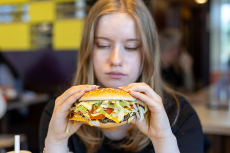Young woman enjoys a large hamburger with fresh ingredients in a casual dining setting during lunchtimeの写真素材