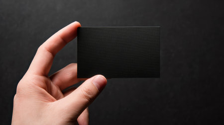 Minimalist black business card held by a hand against a dark textured background in a professional settingの素材