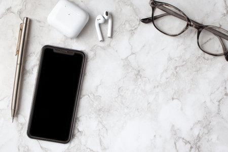 Stylish workspace layout with smartphone, wireless earbuds, glasses, and pen on marble surfaceの素材