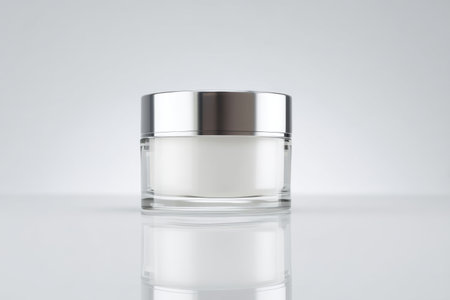 Simple glass jar with silver lid containing white cream on a clean surfaceの素材