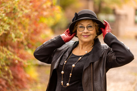 Stylish woman in a black hat and gloves enjoys an autumn stroll amidst colorful foliage in a serene settingの写真素材