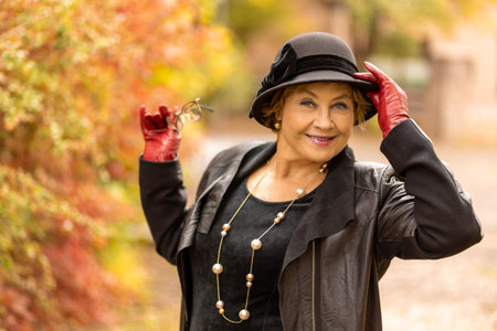 Stylish woman in black leather coat and hat poses with confidence in a colorful autumn settingの写真素材