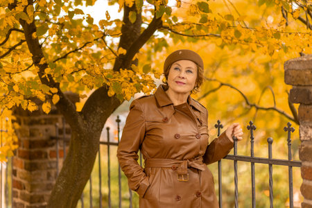 Senior woman in stylish coat poses near a fence surrounded by colorful autumn leaves in a peaceful parkの写真素材