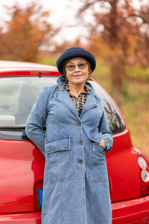 Woman in denim coat poses beside red car in autumn setting during daylight hours