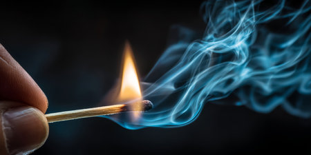 Flame ignites from a matchstick held in hand, creating wisps of smoke in a dimly lit environmentの素材