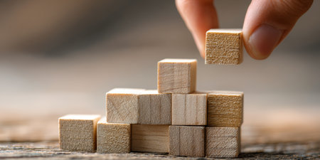 Hand placing wooden block on top of tower of cubes symbolizes growth and balance in a creative workspace during daytimeの素材