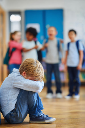 Child sits alone in a school hallway while peers interact, highlighting feelings of isolation and bullying during lunchtimeの素材