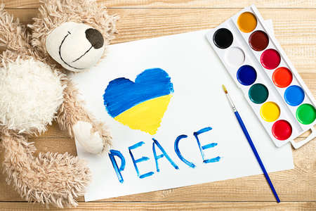 children's drawing with paints with the inscription "peace" and a heart painted in the Ukrainian flag, a teddy bear and watercolors on a wooden tableの写真素材