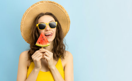 woman in sunglasses and beach hat holds a slice of watermelon on a stick and licks her lips, on a blue backgroundの写真素材