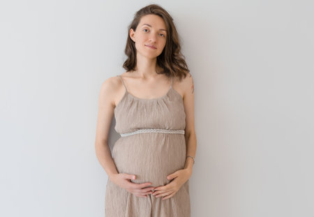 a pregnant woman in a sundress stands against a wall holding her bellyの写真素材