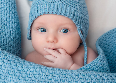 cute shy baby with hand in mouth wearing knitted hat under blanketの写真素材