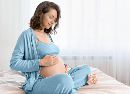 pregnant woman in blue pajamas sits on bed and strokes her belly