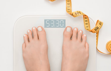 female feet standing on digital scales and measuring tape next to them on light backgroundの写真素材