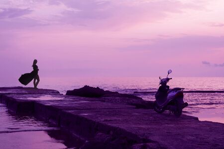 Silhouette of a girl in a fluttering dress and a moped standing on the old sea pier against the background of the sea and sunset. Purple Sea evening landscape. Artistic image. Inspirational scene.の写真素材