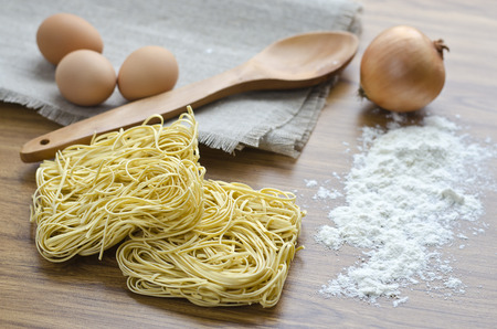 Three eggs and dry egg noodles on the kitchen table and a napkin with a wooden spoon. Ingredients for cooking noodles.の写真素材