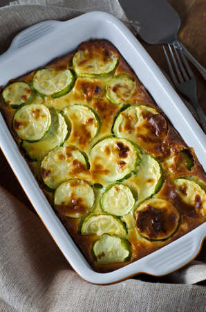 Casserole with zucchini and cheese in a ceramic baking dish on a wooden cutting board.の写真素材