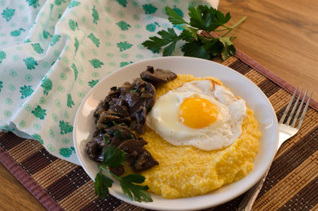 Corn porridge, fried egg, fried mushrooms with onions in a plate on a wicker bamboo napkin. Traditional homemade food. Home tableware on the kitchen table.の写真素材