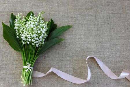 bouquet of white lilies of the valley with green leaves and light ribbon ribbon on linen fabric top view with free space for textの写真素材