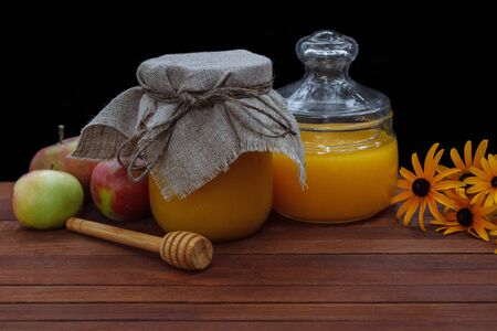 Honey in a glass jar with a linen lid with a bouquet of yellow daisies and apples stands on a brown wooden kitchen table next to a spoon for honey on a dark backgroundの写真素材