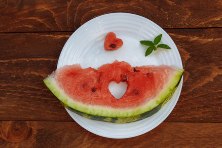 A cut slice of watermelon with seeds on a white plate with a heart cut out of it and a sprig of mint...