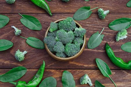 Raw broccoli in a wooden bowl with scattered young spinach leaves and green hot peppers on the kitchen wooden brown table top view close upの写真素材