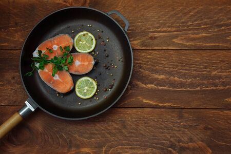 Cut pieces of raw red fish salmon in a cast iron pan with lemon slices spices and green parsley sprigs on a brown wooden background top view with an empty space for textの写真素材