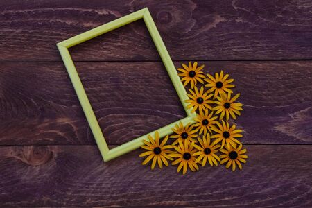 Yellow empty photo frame with yellow Daisy flowers laid out in the corner top view with free space for lettering and text on a brown wooden backgroundの写真素材