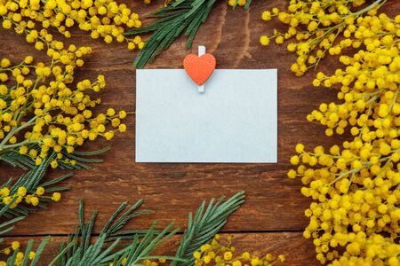 A frame of yellow Mimosa flowers with green leaves with an empty paper card and a paper clip with a red heart on a brown wooden backgroundの写真素材