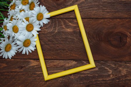Empty yellow plastic photo frame with a bouquet of white daisies on a brown wooden background top...