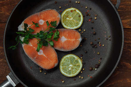 Slices of pink salmon in a black cast iron pan with a bunch of green parsley, lemon and spices top view on a koichnevom wooden background, the concept of cooking lunch, restaurant, healthy food, omegaの写真素材